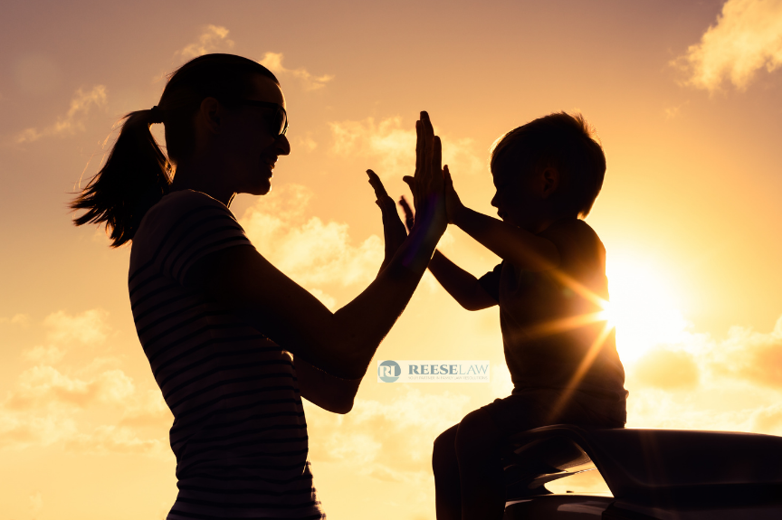Silhouette of a parent and young child giving each other a high-five while sitting outdoors at sunset, symbolizing a positive co-parenting relationship and shared moments with children after divorce. ReeseLaw logo appears near the center of the image.